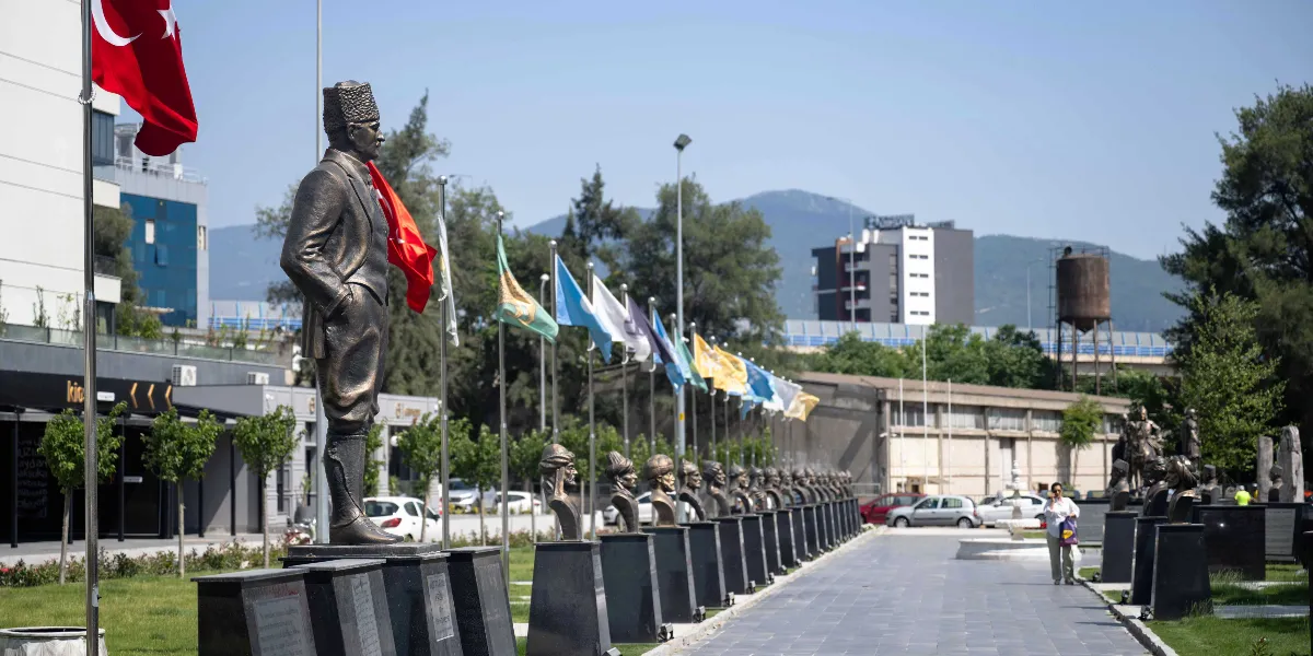 Abide-i Hürriyet Meydanı'nda, ön planda Mustafa Kemal Atatürk'ün tam boy heykeli ve arkasında sıra halinde dizilmiş tarihi şahsiyetlere ait büstler yer alıyor. Büstlerin arkasında farklı renklerde bayraklar dalgalanırken, arka planda ağaçlar, şehir yapıları ve dağ manzarası görülüyor. Yürüyüş yolunda bir kişi yürüyor ve çevrede düzenlenmiş yeşil alanlar bulunuyor.