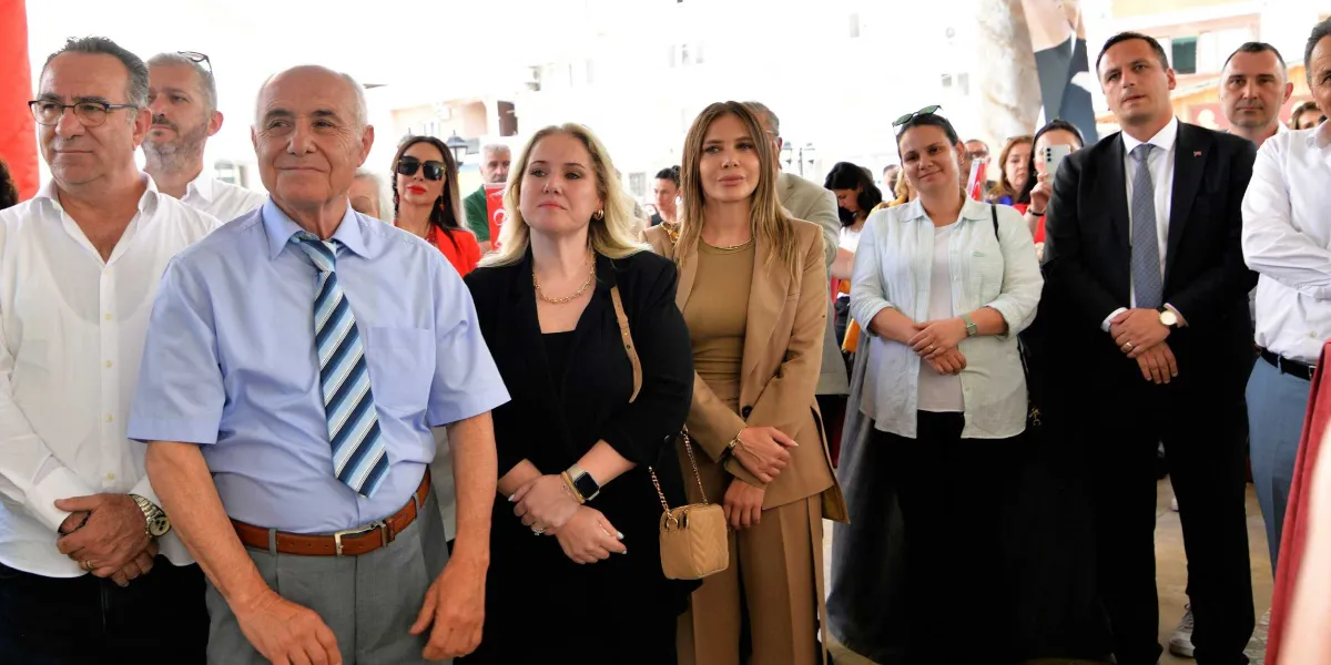 Açık havada düzenlenen bir törende, aralarında Bornova Belediye Başkanı Ömer Eşki'nin de bulunduğu bir grup insan yan yana durarak dikkatle bir noktaya bakıyor. Ön planda, açık mavi gömlek ve çizgili kravat takan, gümüş saçlı bir beyefendi ile yanında siyah ceketli sarışın bir kadın ve bej renkli takım elbise giyen bir kadın yer alıyor. Arka planda ise takım elbiseli erkekler ve çeşitli kıyafetler içindeki diğer katılımcılar bulunuyor.