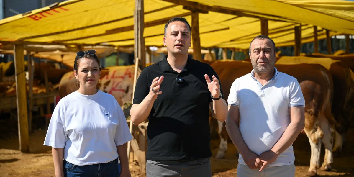 Sarı tenteli bir kurban satış alanında, beyaz tişörtlü bir kadın, siyah polo yaka tişörtlü konuşan bir erkek ve beyaz polo yaka tişörtlü bir erkek yan yana duruyor. Arkalarında kurbanlık büyükbaş hayvanlar ve ahşap direklerle desteklenmiş çadır yapısı görülüyor.