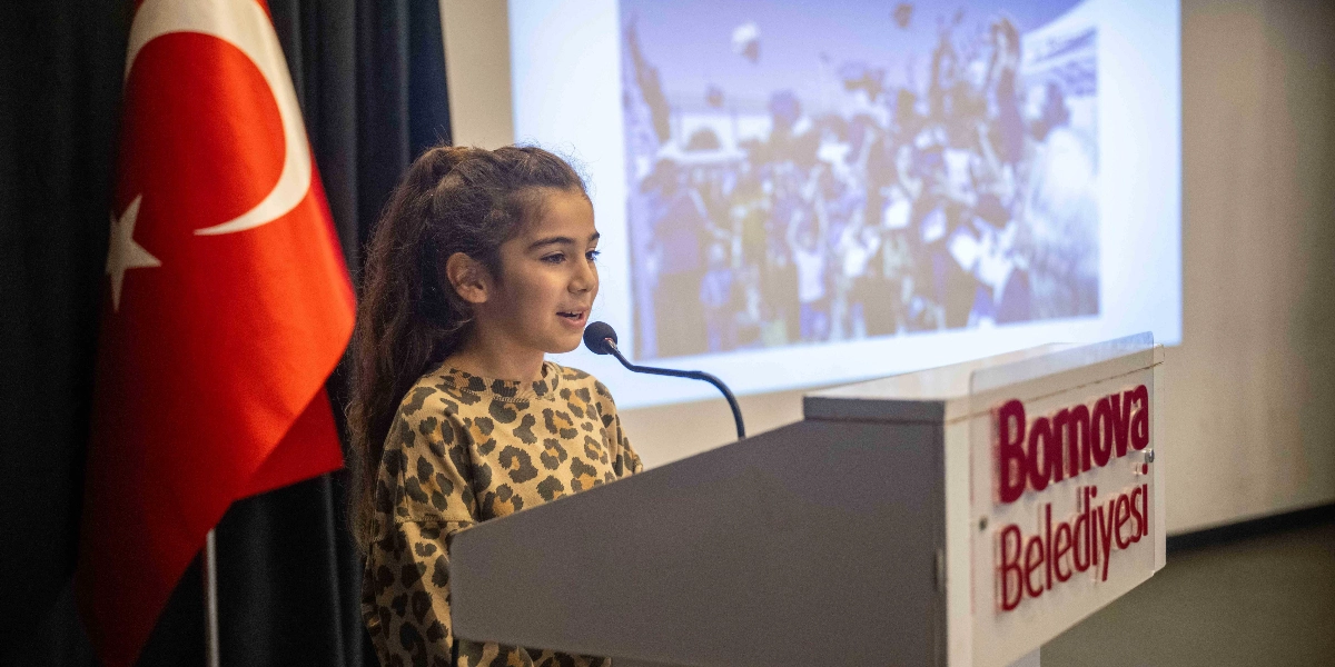 Leopar desenli bluz giymiş genç bir kız, üzerinde Bornova Belediyesi yazan bir kürsüde mikrofon başında konuşma yapıyor. Arka planda Türk bayrağı ve üzerinde kalabalık bir grubun yer aldığı yansıtılmış bir görsel bulunuyor.