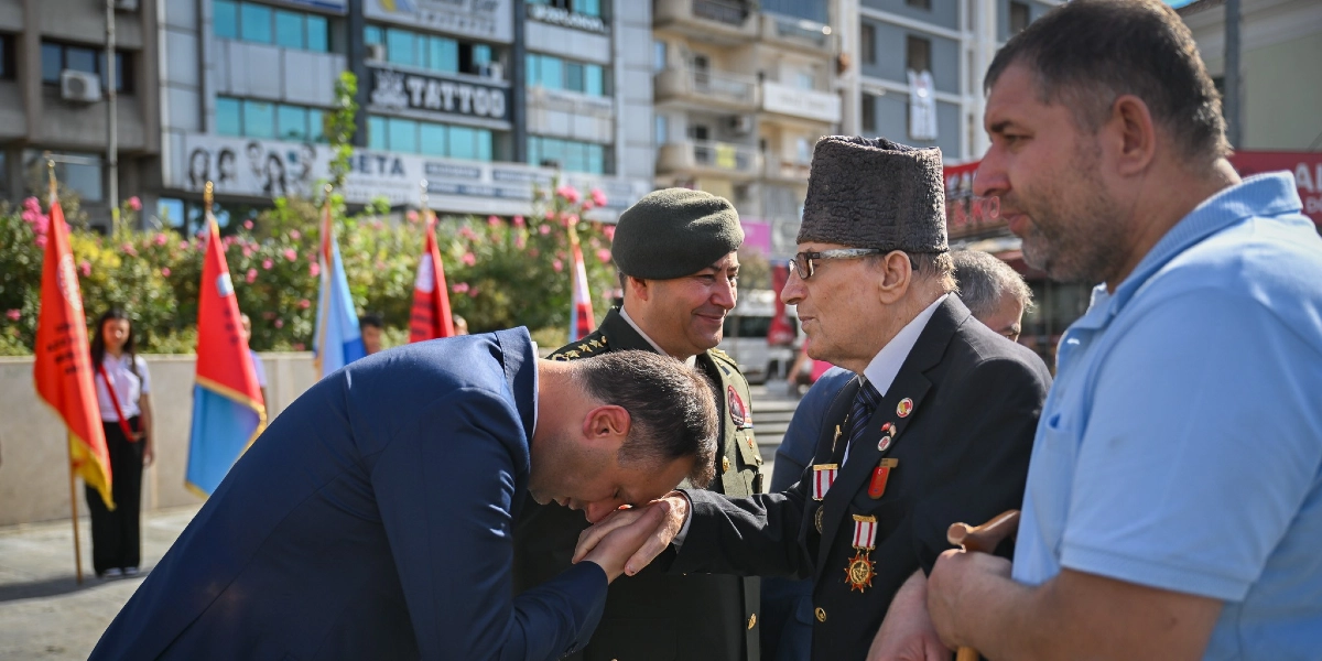 Lacivert takım elbiseli bir erkek, 19 Eylül Gaziler Günü töreninde madalyalı yaşlı bir gazinin elini öperek saygısını gösteriyor. Gazinin yanında askeri üniformalı bir subay ve sağ tarafta mavi tişörtlü bir vatandaş yer alıyor. Arka planda ise tören alanındaki bayraklar ve şehir merkezi binaları görülüyor.