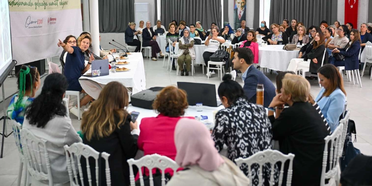 Geniş bir salonda düzenlenen toplantıda, beyaz örtülü yuvarlak masaların etrafında toplanmış çok sayıda kadın katılımcı yer alıyor. Salonun ön tarafında, üzerinde dizüstü bilgisayarların bulunduğu uzun bir masada oturan bir kadın, projeksiyon ekranına doğru işaret ederek konuşma yapıyor. Arka planda duvarda Atatürk posteri ve Türk bayrağı asılı bulunurken, katılımcılar dikkatle dinliyor. Masaların üzerinde yiyecek ve içecekler yer alıyor.