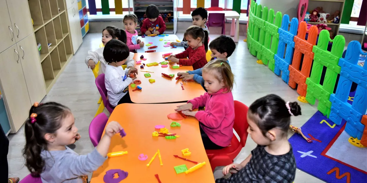 Renkli plastik çitlerle çevrili bir oyun odasında, turuncu masaların etrafında oturan çocuklar oyun hamurlarıyla etkinlik yapıyor. Çocuklar masaların üzerindeki çeşitli şekillerdeki hamur kalıpları ve merdanelerle oynarken, odanın bir köşesinde oyuncak rafları ve üzerinde harflerin bulunduğu renkli bir halı yer alıyor.
