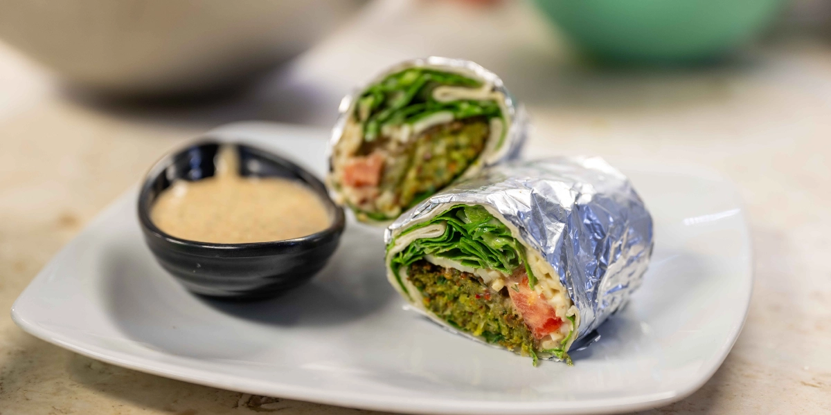 Alüminyum folyoya sarılmış, içi falafel, taze yeşillikler ve domates dilimleriyle dolu ikiye bölünmüş bir dürüm, yanında küçük siyah bir kapta sos ile birlikte beyaz bir tabak üzerinde servis ediliyor.