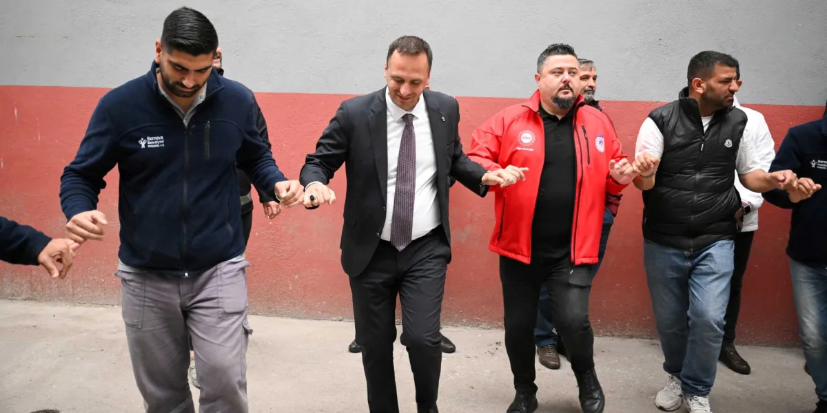 Bornova Belediye Başkanı Ömer Eşki, üzerinde takım elbise ve kravatıyla, 1 Mayıs kutlamaları kapsamında işçilerle el ele tutuşarak halay çekiyor. Eşki'nin sağında Bornova Belediyesi logolu lacivert polar giyen bir çalışan, solunda ise kırmızı ceketli bir kişi yer alıyor. Arka planda gri ve kırmızı renkli bir duvar bulunuyor.
