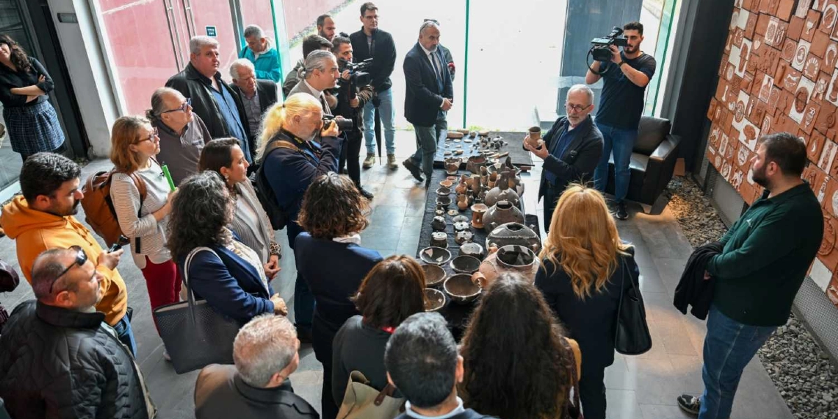 İzmir'deki bir müze veya sergi alanında, arkeolojik buluntuların sergilendiği uzun bir masanın etrafında toplanmış bir grup insan. Gözlüklü bir uzman, elinde tuttuğu antik bir çömleği katılımcılara göstererek açıklamalarda bulunuyor. Masanın üzerinde çok sayıda farklı boyut ve şekilde tarihi seramik kaplar ve objeler yer alıyor. Etrafta fotoğraf çeken basın mensupları ve dikkatle dinleyen ziyaretçiler bulunurken, sağ taraftaki duvarda ise pişmiş topraktan yapılmış rölyef panolar sergileniyor.