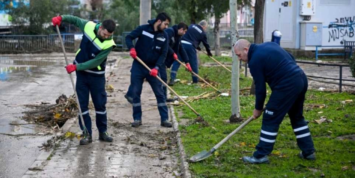 Belediye üniformalı beş işçi, sel sonrası biriken çamur ve atıkları temizlemek için ellerinde küreklerle kaldırım ve yeşil alanda çalışma yapıyor.