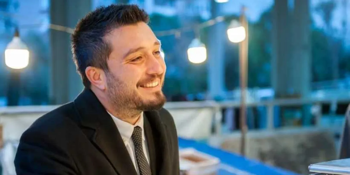 Koyu renkli ceket, beyaz gömlek ve desenli kravat giymiş, hafif sakallı, gülümseyen genç bir adam. Arka planda akşam saatlerinde dış mekanda asılı duran dekoratif aydınlatmalar bulunuyor.