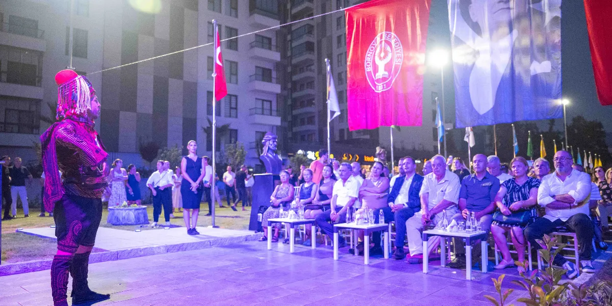 Gece vakti açık havada düzenlenen bir etkinlikte, geleneksel Osmanlı kıyafetleri giymiş bir kişi ayakta durarak izleyicilere hitap ediyor. Etkinlik alanında Bornova Belediyesi bayrağı ve üzerinde Atatürk portresi bulunan büyük bir afiş asılı. Arka planda modern apartmanlar ve aydınlatılmış bir Atatürk büstü yer alırken, ön tarafta sandalyelerde oturan kalabalık bir izleyici grubu etkinliği takip ediyor.