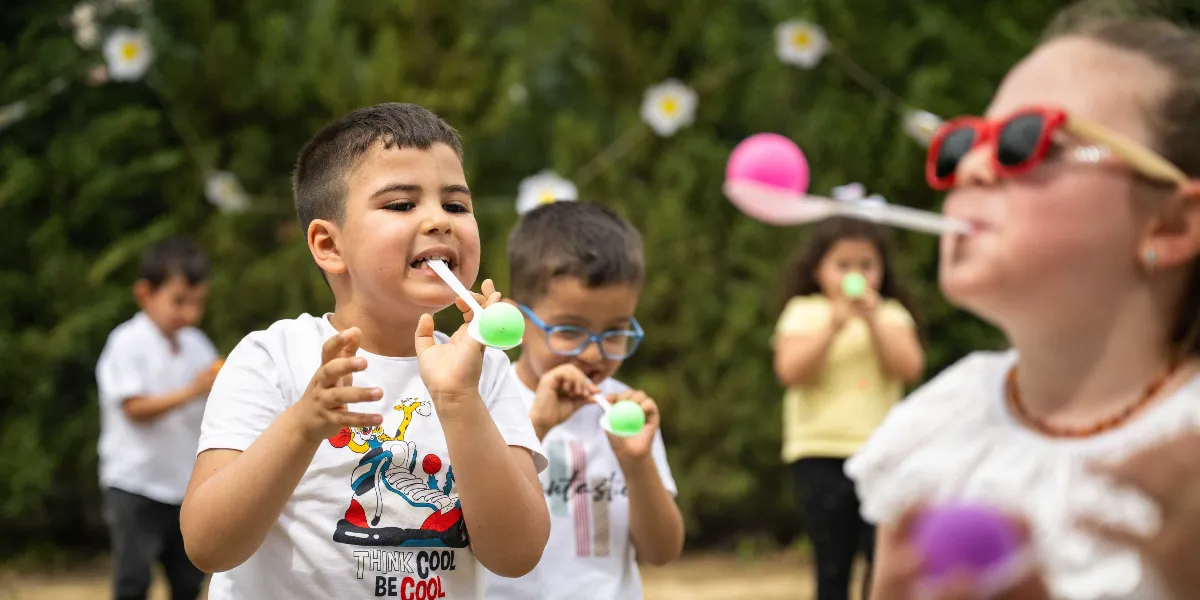 Açık havada, yeşil bir arka plan önünde beyaz tişörtlü çocuklar, ağızlarında tuttukları plastik kaşıkların üzerinde renkli topları düşürmeden taşıma yarışı yapıyor. Ön planda odak noktasında olan bir erkek çocuk ve kırmızı güneş gözlüklü bir kız çocuğu dikkatle dengede durmaya çalışan toplara odaklanmış durumda; arka planda ise oyuna devam eden diğer çocuklar yer alıyor.
