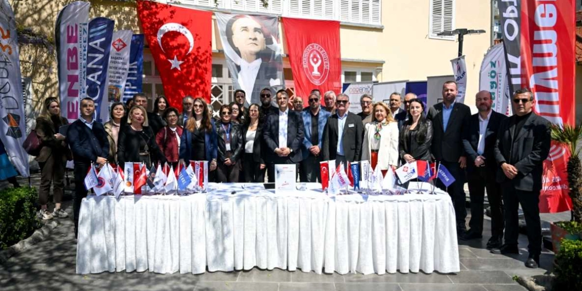 Açık havada, beyaz örtülü uzun bir masanın arkasında yan yana duran, aralarında sağlık sektörü temsilcilerinin de bulunduğu kalabalık bir grup insan. Arka planda Türk bayrağı, Mustafa Kemal Atatürk'ün portresi ve Bornova Belediyesi'nin flaması asılıdır. Grubun etrafında çeşitli kurum ve kuruluşlara ait dikey tanıtım bayrakları yer alırken, masanın üzerinde de küçük masa bayrakları ve bir protokol dosyası bulunmaktadır.