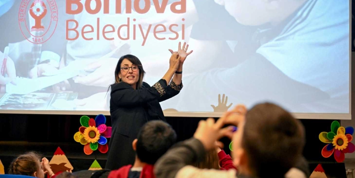 Bornova Belediyesi logosunun bulunduğu bir projeksiyon perdesinin önünde, gözlüklü bir kadın eğitmen, öğrencilere ellerini kullanarak uygulamalı bir sağlık eğitimi veriyor. Eğitmen, el hareketleriyle bir konuyu anlatırken, ön tarafta oturan öğrenciler de onu dikkatle izliyor. Sahne dekorunda renkli çiçek figürleri ve kalem görselleri yer alıyor.
