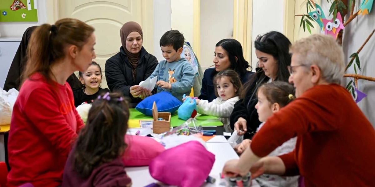 Bir sınıf ortamında, masanın etrafında toplanmış kadınlar ve çocuklar el işi etkinliği yapıyor. Yetişkinler ve çocuklar birlikte kumaş parçaları, yastıklar ve dikiş malzemeleriyle uğraşırken, ortamda neşeli bir atmosfer hakim. Arka planda duvarda baykuş figürlü süslemeler ve çocuklara yönelik dekoratif öğeler yer alıyor.