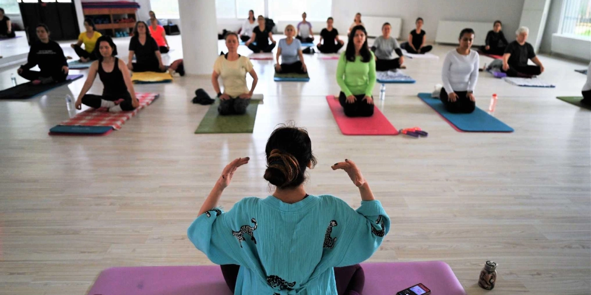 Aydınlık ve geniş bir spor salonunda, yoga eğitmeninin arkasından çekilmiş geniş açılı karede, eğitmen matının üzerinde oturmuş ellerini havaya kaldırmış şekilde dersi yönetiyor. Karşısında, farklı renklerdeki yoga matları üzerinde bağdaş kurmuş veya diz çökmüş şekilde oturan, çeşitli yaş gruplarından kadınlar dikkatle eğitmeni izliyor. Salonun zemininde ahşap parke kaplama bulunuyor ve katılımcıların önlerinde su şişeleri ve kişisel eşyaları yer alıyor.