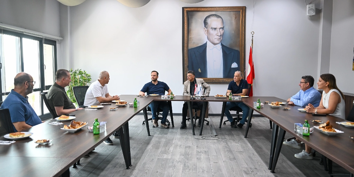 U-şeklindeki büyük bir toplantı masasının etrafında oturan sekiz kişilik bir grup, sohbet ederken görülüyor. Masanın üzerinde tabaklarda börekler ve içecekler bulunuyor. Arka duvarda büyük bir Mustafa Kemal Atatürk portresi ve yanında Türk bayrağı yer alıyor. Modern bir ofis ortamındaki bu toplantıda katılımcılar günlük kıyafetler giyiyor.