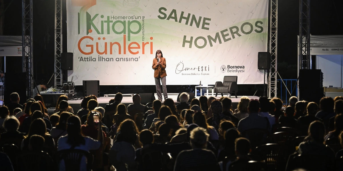 Gece vakti açık havada düzenlenen bir etkinlikte, sahnede mikrofonla konuşma yapan bir kadın ve onu izleyen kalabalık bir dinleyici grubu görülüyor. Sahne arkasındaki büyük pankartta 