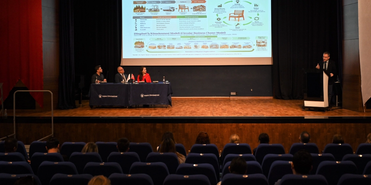 Yaşar Üniversitesi'nde düzenlenen bir etkinlikte, sahnedeki kürsüde konuşma yapan bir kişi ve masada oturan üç panelist yer alıyor. Sahnenin arka planındaki dev ekranda, döngüsel ekonomi ve döngüsel iş kümelenmesi modelini açıklayan şemalar, grafikler ve metinler bulunuyor. İzleyiciler, sahnenin önündeki koltuklarda oturarak sunumu takip ediyor.