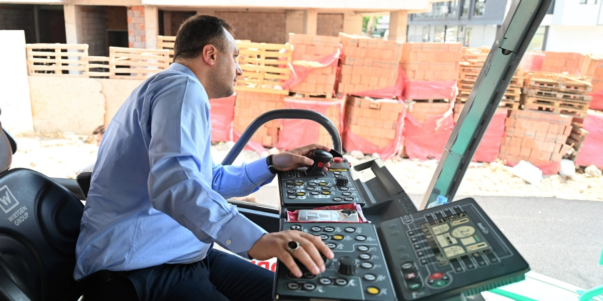 Mavi gömlekli bir erkek, Wirtgen Group marka bir iş makinesinin operatör koltuğunda oturarak kontrol panelini yönetiyor. Arka planda inşaat alanında üst üste dizilmiş tuğla paletleri ve devam eden bir yapı çalışması görülüyor.