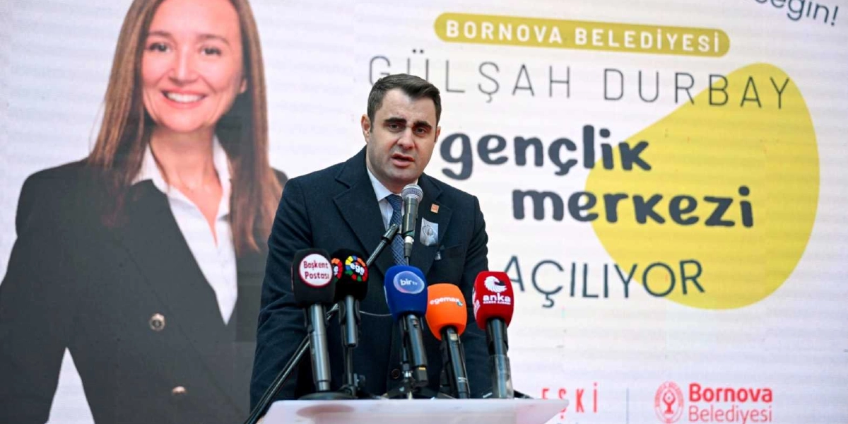 Koyu renk ceket ve kravat giymiş bir erkek, önünde mikrofonlarla bir kürsüde konuşma yapıyor. Arka planda Bornova Belediyesi Gülşah Durbay Gençlik Merkezi'nin açılışını duyuran bir afiş yer alıyor; afişin sol tarafında Gülşah Durbay'ın gülümseyen bir fotoğrafı, sağ tarafında ise büyük puntolarla 