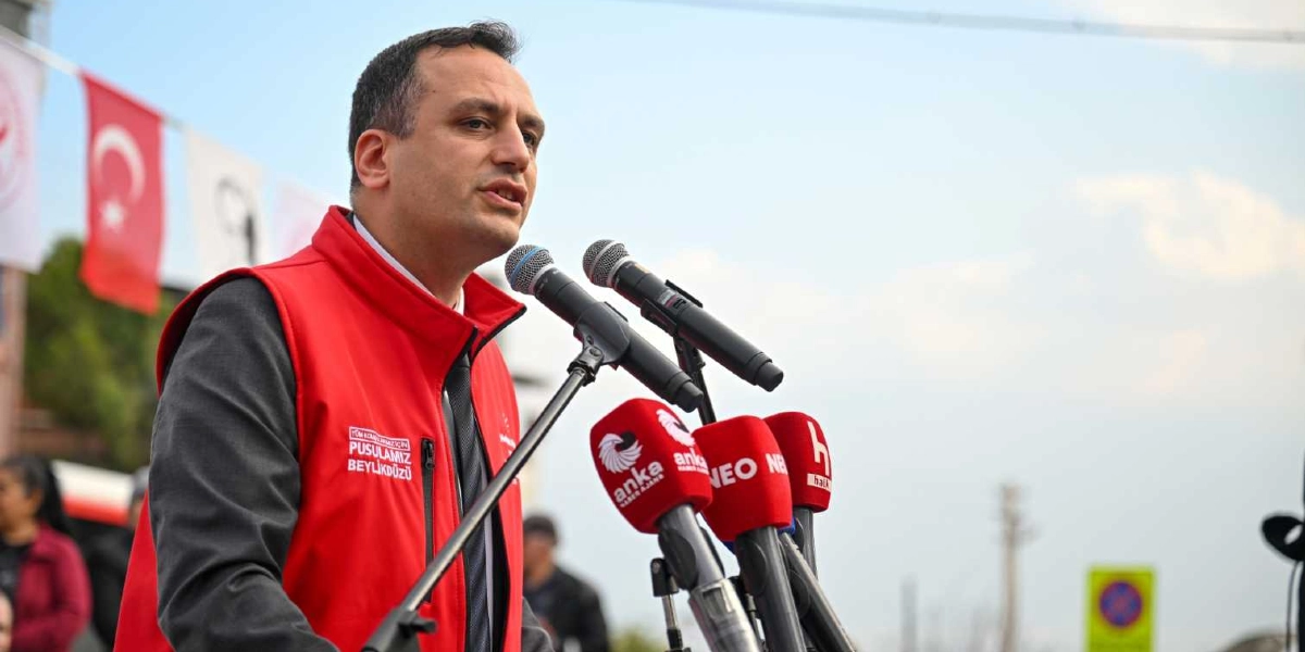 Koyu renk ceket ve kravat üzerine kırmızı bir yelek giymiş bir erkek, açık havada kürsüde konuşma yapıyor. Yeleğinin üzerinde 
