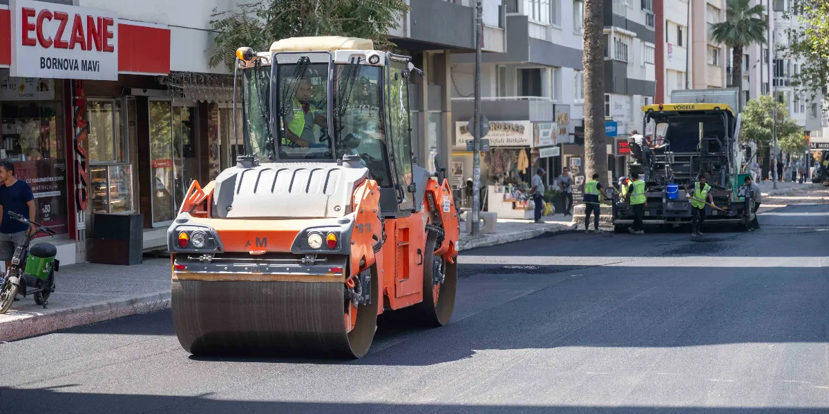Bornova'da bir cadde üzerinde asfalt yenileme çalışması yapılıyor. Ön planda turuncu renkli bir silindir operatörüyle birlikte ilerlerken, arka planda Vögele marka bir asfalt serici makine ve yelekli işçiler çalışmaya devam ediyor. Yolun kenarında 