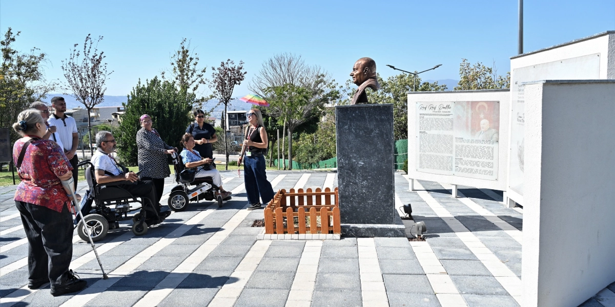 Açık havada, park benzeri bir alanda Rauf Raif Denktaş'ın büstü ve hayatını anlatan bilgilendirme panosunun önünde duran bir grup insan. Grupta tekerlekli sandalye kullanan bireyler ve onlara eşlik eden bir rehber bulunuyor. Rehber, elinde renkli bir şemsiye tutarak grubu bilgilendiriyor. Zemin, gri ve beyaz taşlarla döşenmiş, güneşli bir gün.