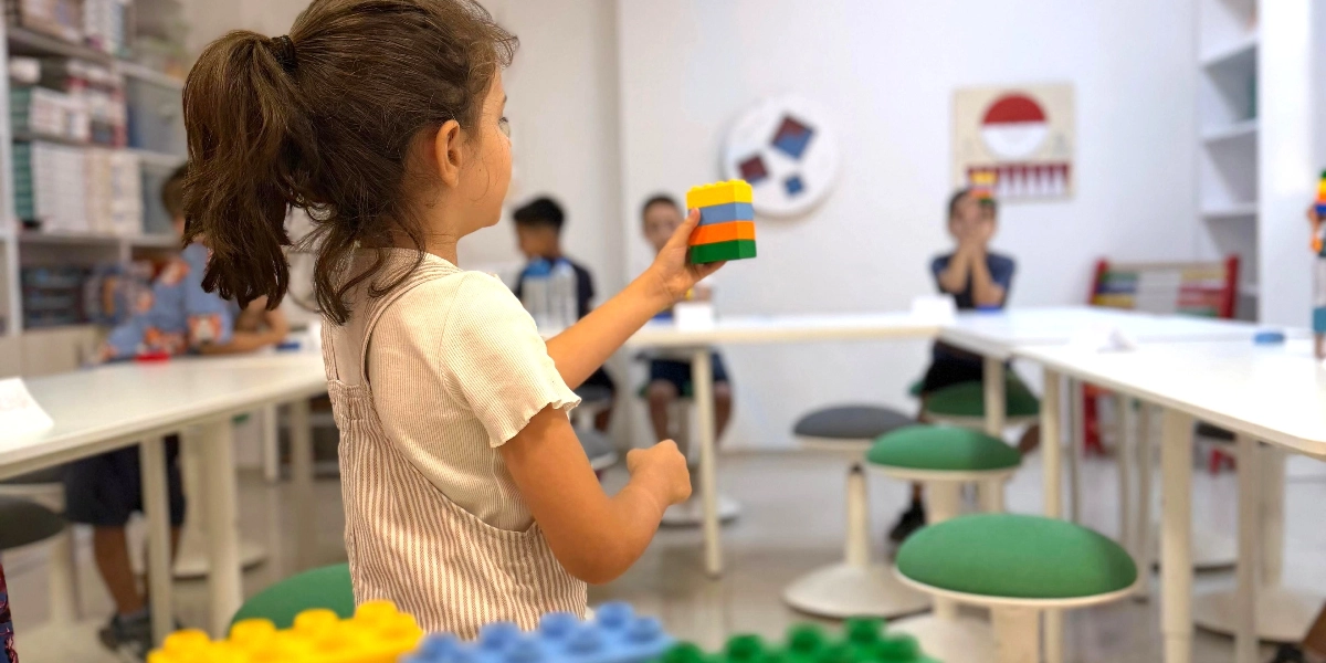 At kuyruğu saçlı küçük bir kız çocuğu, elinde tuttuğu üst üste dizilmiş dört farklı renkteki yapı bloğunu sınıf arkadaşlarına gösteriyor. Aydınlık bir sınıfta, beyaz masaların etrafında yeşil taburelerde oturan diğer çocuklar etkinliği izliyor. Ön planda, odak dışı kalmış renkli yapı blokları yer alıyor.