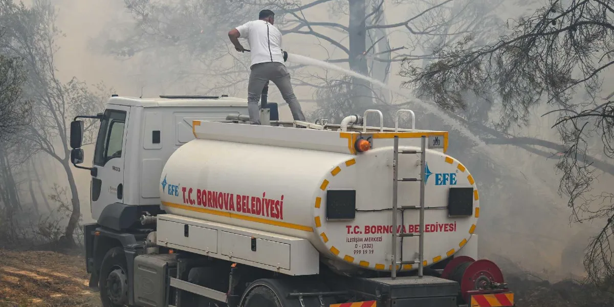 Üzerinde T.C. Bornova Belediyesi yazılı beyaz bir arazözün tepesinde duran bir görevli, hortumla ormanlık alandaki yangına su sıkarak müdahale ediyor. Arka planda dumanlar arasında ağaçlar görülüyor.