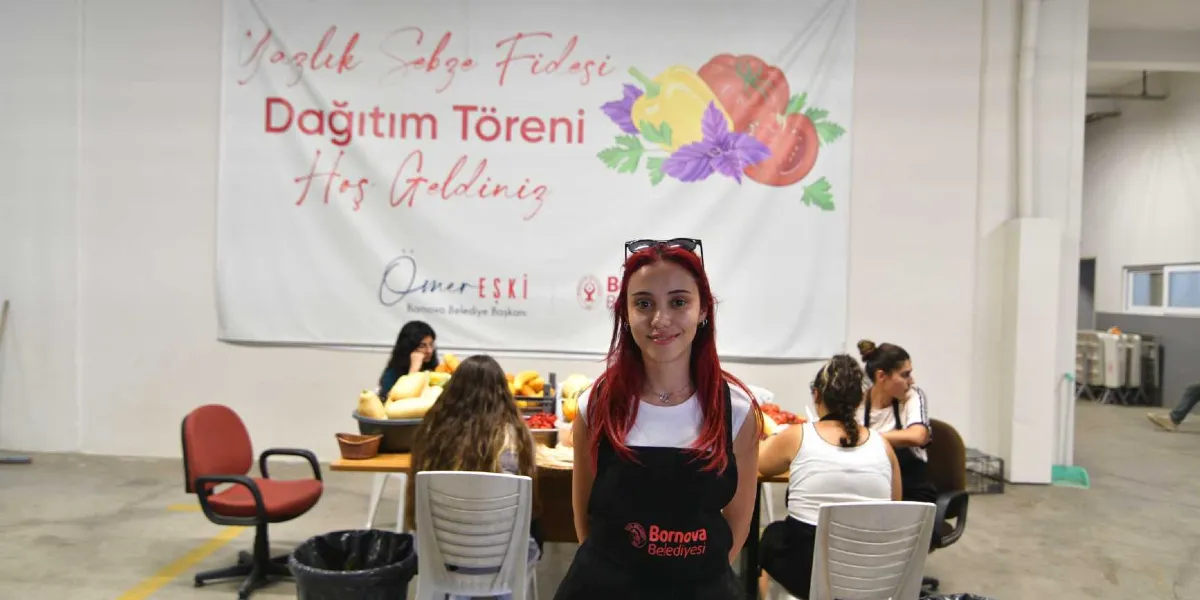 Kızıl saçlı genç bir kadın, üzerinde Bornova Belediyesi logolu siyah bir önlükle kameraya gülümseyerek bakıyor. Arkasında, üzerinde 