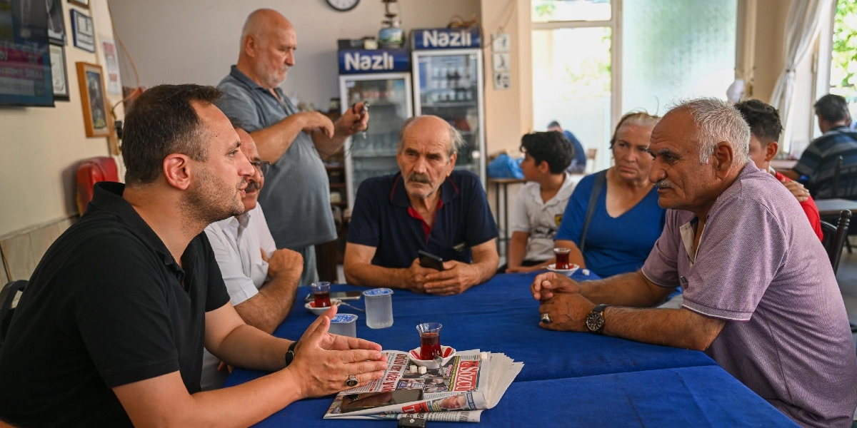 Belediye başkanı, Ümit Mahallesi'ndeki bir kahvehanede mavi örtülü bir masanın etrafında oturan vatandaşlarla sohbet ediyor. Masanın üzerinde çay bardakları ve bir gazete bulunurken, arka planda ayakta duran bir kişi ve diğer masalarda oturan insanlar yer alıyor.