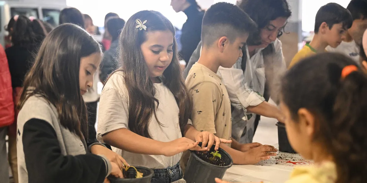 Bir masa etrafında toplanmış çocuklar, önlerindeki saksılara toprak ekleyerek bitki dikiyor. Odak noktasında, saçında kelebek şeklinde bir toka bulunan kız çocuğu dikkatle saksıdaki toprağı düzeltiyor; yanında diğer çocuklar ve onlara rehberlik eden bir yetişkin bulunuyor.