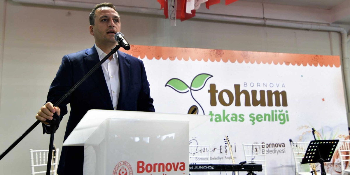 Koyu renk ceket ve beyaz gömlek giymiş bir yetkili, üzerinde Bornova Belediyesi logosu bulunan bir kürsüde mikrofon başında konuşma yapıyor. Arka planda 