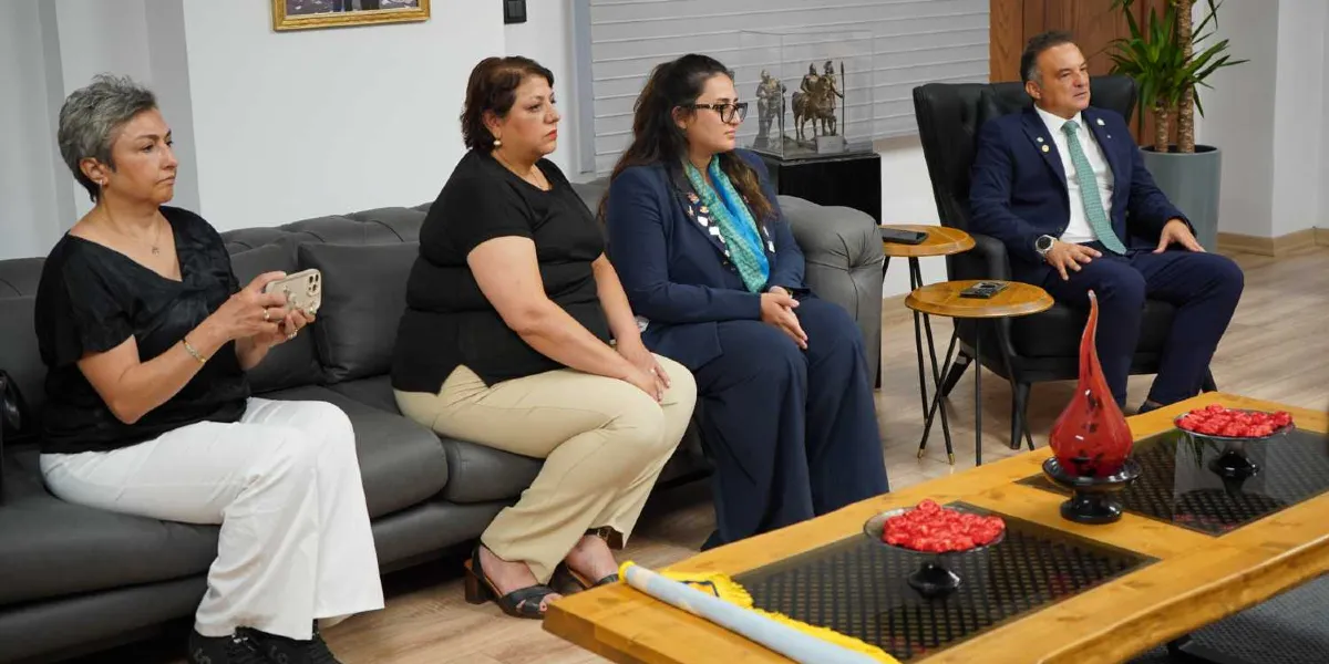 Modern bir ofis ortamında, gri bir koltukta yan yana oturan üç kadın ve tekli koltukta oturan bir erkekten oluşan grup. Soldaki kadın elinde cep telefonu tutarken, yanındaki iki kadın ve sağ taraftaki takım elbiseli erkek karşıya doğru bakıyor. Önlerinde ahşap bir orta sehpa, üzerinde kırmızı dekoratif objeler ve bir rulo bulunuyor. Arka planda ise cam bir fanus içinde heykelcik ve yeşil bir bitki yer alıyor.
