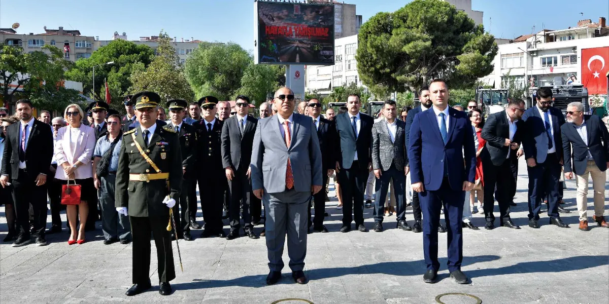 Açık havada, bir meydanda düzenlenen resmi törende yan yana dizilmiş, takım elbiseli ve askeri üniformalı protokol üyeleri. En önde, ortada gri takım elbiseli bir beyefendi, sağında lacivert takım elbiseli bir beyefendi ve solunda askeri üniformalı bir subay yer alıyor. Arka planda binalar, ağaçlar, üzerinde 
