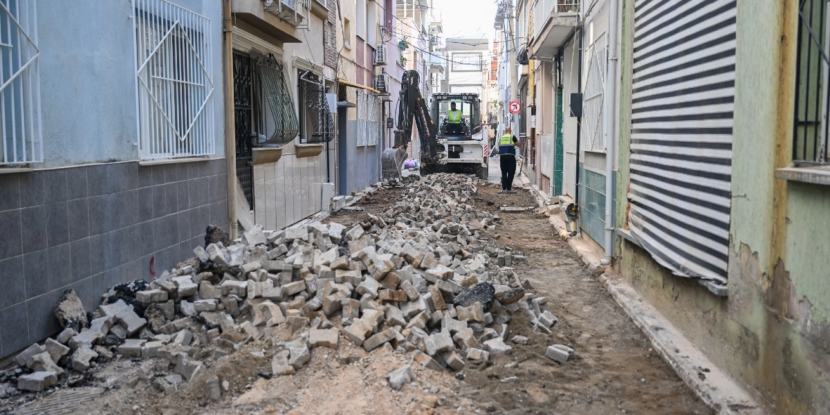 Bornova'da dar bir sokakta devam eden yol yenileme çalışmaları kapsamında, sökülmüş parke taşlarının oluşturduğu yığınlar ve arkada çalışan bir iş makinesi görülüyor. İş makinesini kullanan operatör ve yanında duran görevli, sarı reflektörlü yelekleriyle çalışmalara eşlik ediyor.