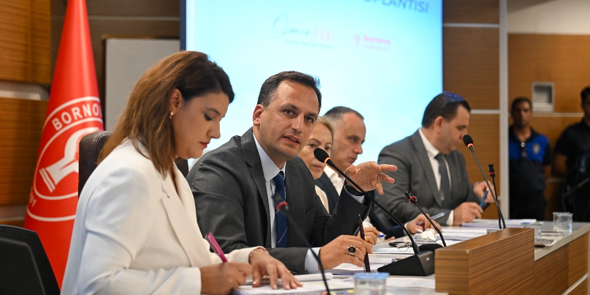 Bornova Belediye Başkanı Ömer Eşki, bir toplantı masasında mikrofon önünde konuşma yaparken görülüyor. Yanında beyaz ceketli bir kadın not alırken, masanın devamında diğer katılımcılar yer alıyor. Arka planda Bornova Belediyesi'nin kırmızı bayrağı ve üzerinde toplantı başlığı bulunan bir projeksiyon ekranı bulunuyor.
