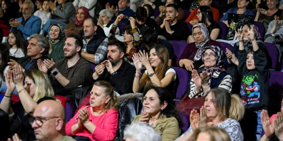Kapalı bir salonda, mor koltuklarda oturan ve alkışlayarak bir etkinliği izleyen, farklı yaş gruplarından kadın ve erkeklerin oluşturduğu kalabalık bir izleyici grubu. Ön planda, pembe bluzlu bir kadın ve yanında oturan, gözlüklü ve saçları başörtülü yaşlı bir kadın dikkat çekerken, sağ tarafta ellerini havaya kaldırarak neşeyle alkışlayan küçük bir çocuk görülüyor. İzleyicilerin çoğu sahneye doğru bakarak gülümseyen ifadelerle etkinliğe katılım gösteriyor.