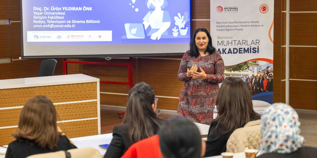 Doç. Dr. Ürün Yıldıran Önk, Bornova Muhtarlar Akademisi kapsamında düzenlenen bir eğitimde katılımcılara sunum yapıyor. Arka planda üzerinde konuşmacının bilgilerinin yer aldığı bir projeksiyon perdesi ve 