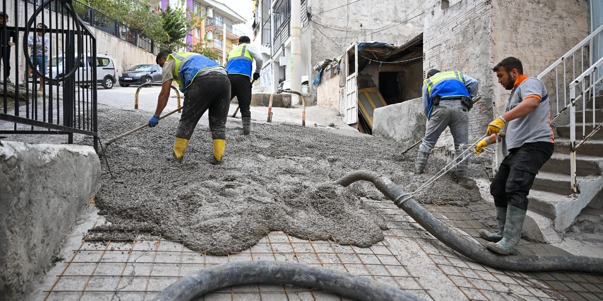 Belediye işçileri, Bornova'da eğimli bir sokakta beton dökme ve yol düzenleme çalışması yapıyor. İşçilerden bazıları ellerindeki tırmık ve küreklerle taze betonu düzeltirken, bir işçi beton pompası hortumunu yönlendiriyor. Çalışma alanında zemin üzerinde çelik hasır demirler görülüyor ve arka planda binalar ile park halindeki araçlar yer alıyor.