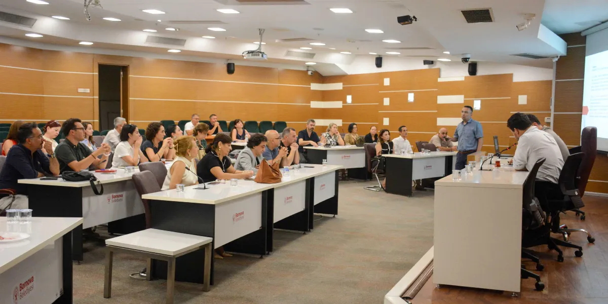 Bornova Belediyesi'ne ait modern bir toplantı salonunda, katılımcıların sıralar halinde oturduğu ve bir konuşmacının ayakta sunum yaptığı eğitim veya bilgilendirme toplantısı. Salonun ön tarafında projeksiyon ekranı, masaların üzerinde Bornova Belediyesi logoları ve katılımcıların önünde su bardakları yer alıyor.