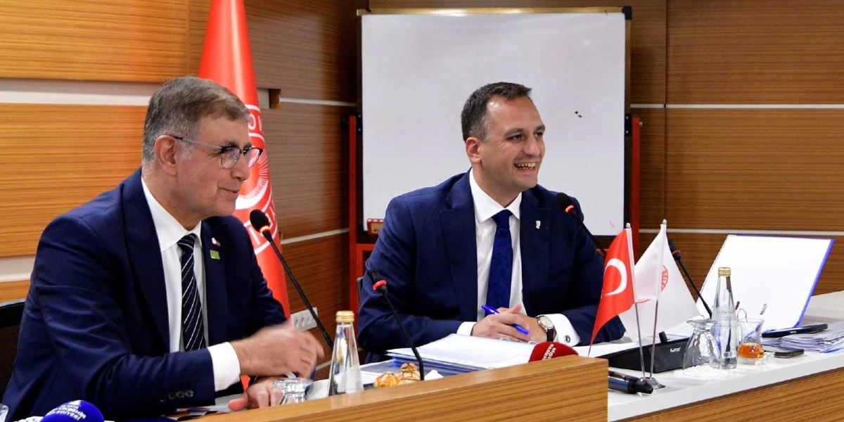 Takım elbiseli ve kravatlı iki erkek, bir toplantı masasında yan yana oturarak gülümseyerek sohbet ediyor. Masanın üzerinde mikrofonlar, Türk bayrağı, belediye amblemli bir bayrak, su şişeleri ve dosyalar bulunuyor. Arka planda ahşap panelli bir duvar ve beyaz bir yazı tahtası yer alıyor.