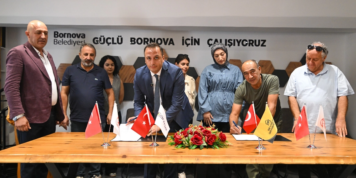 Bornova Belediyesi yetkilileri ve iş birliği yapılan kurum temsilcileri, üzerinde Türk bayrakları ve kurumsal logoların bulunduğu ahşap bir masada protokol imzalıyor. Arka planda duvarda Bornova Belediyesi logosu ve 