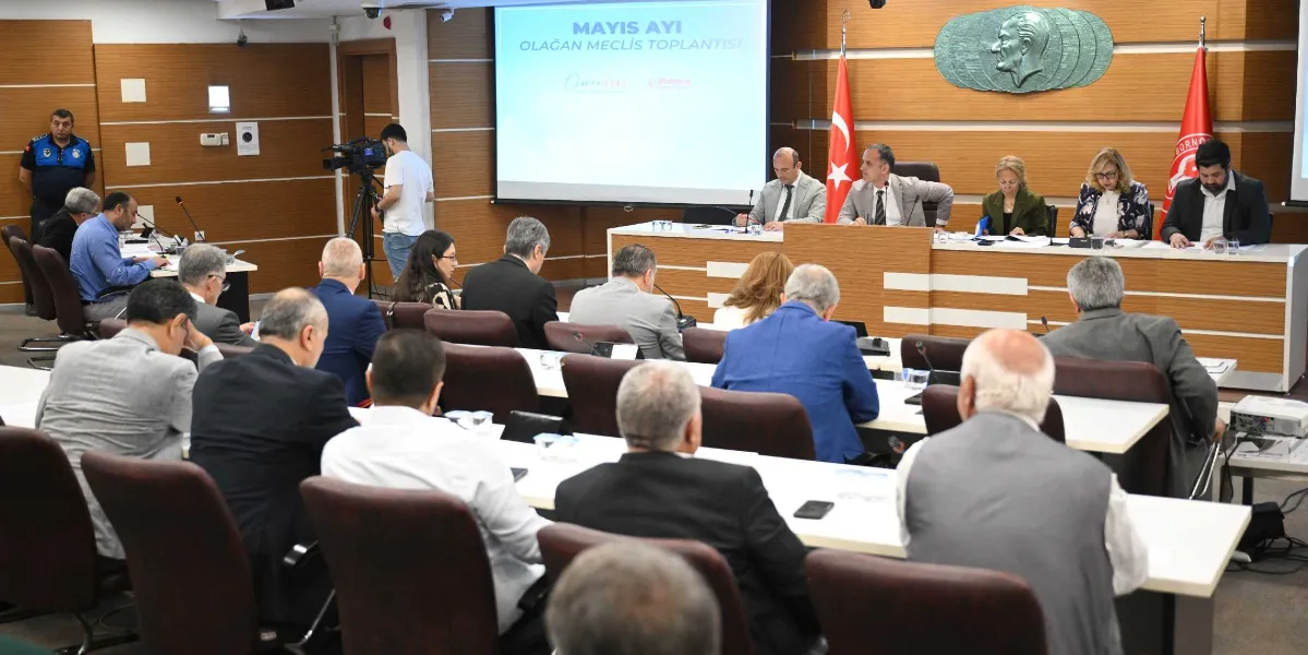 Bornova Belediyesi meclis salonunda gerçekleşen toplantıda, katılımcılar sıralarda oturarak gündemi takip ediyor. Salonun ön kısmındaki kürsüde meclis başkanlık divanı üyeleri yer alırken, arkadaki projeksiyon ekranında 