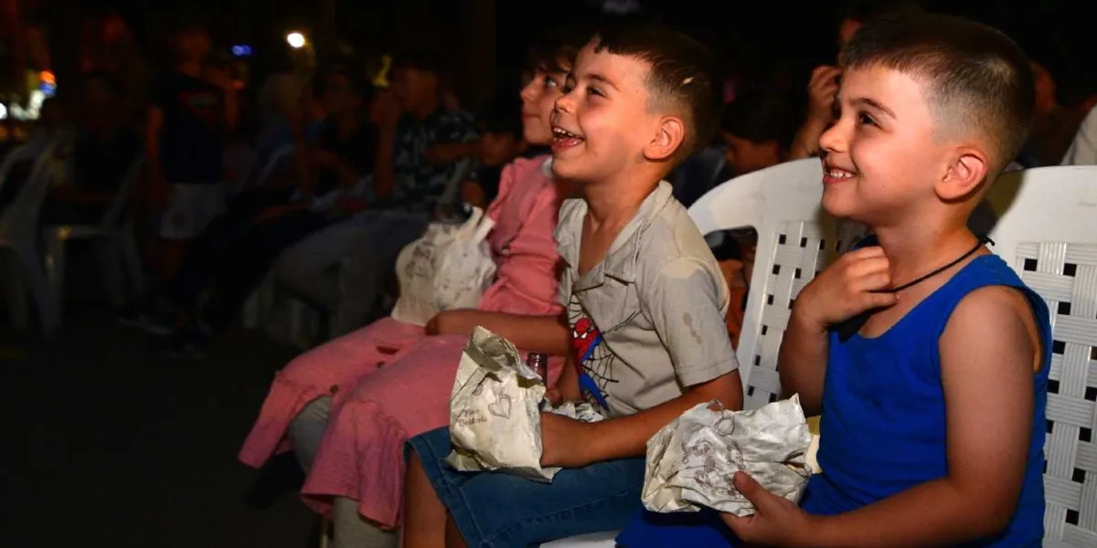 Açık havada düzenlenen bir sinema etkinliğinde, beyaz plastik sandalyelerde yan yana oturarak keyifle film izleyen ve ellerinde patlamış mısır paketleri tutan neşeli çocuklar.