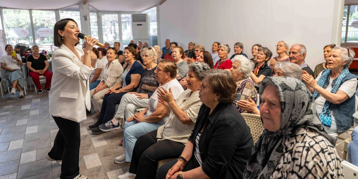 Beyaz ceketli bir kadın, elinde mikrofonla ayakta şarkı söylerken, karşısında oturan yaşlılardan oluşan bir grup dinleyici ona eşlik ediyor ve alkışlıyor. Aydınlık ve geniş bir salonda gerçekleşen etkinlikte, katılımcılar keyifli bir atmosferde bir arada bulunuyor.