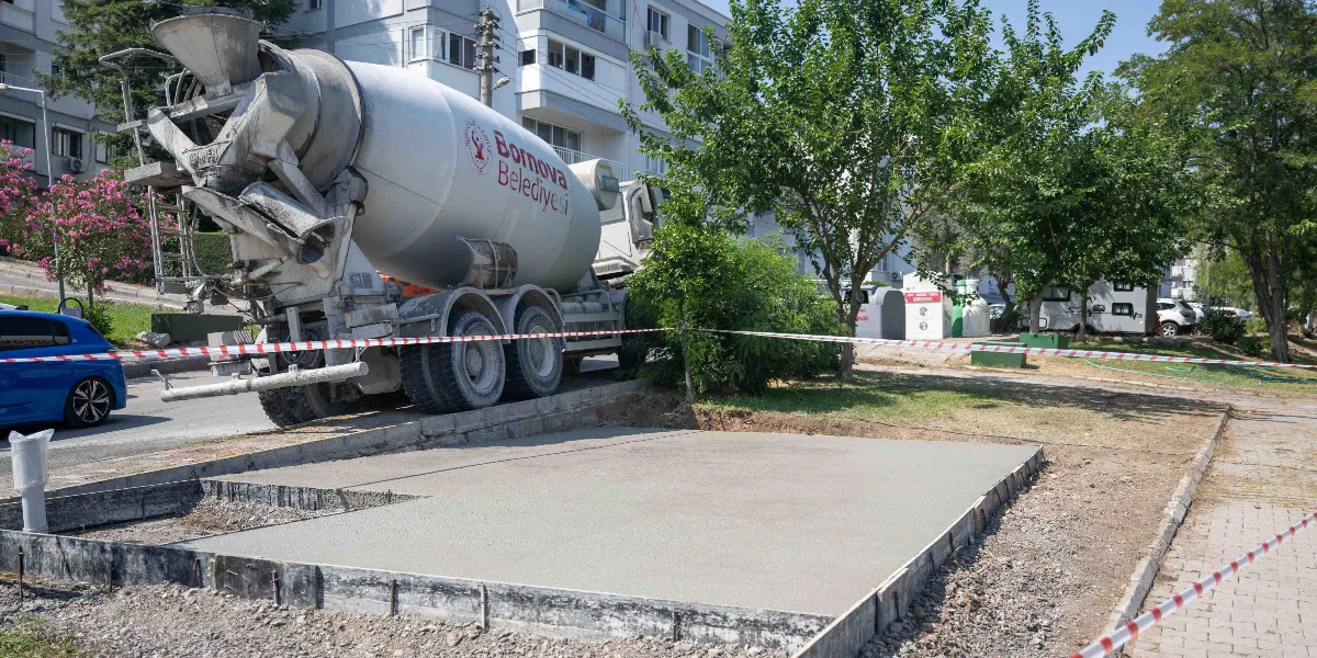 Bornova Belediyesi'ne ait bir beton mikseri, Evka 3 mahallesinde yeni dökülmüş beton bir zemin çalışmasının yanında park halinde duruyor. Çalışma alanı kırmızı beyaz güvenlik şeridiyle çevrilmiş olup, arka planda apartmanlar ve yeşil ağaçlar görülüyor.