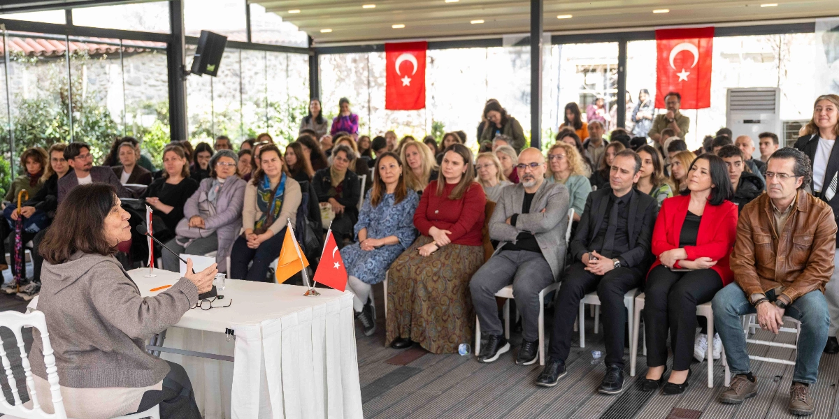 Yazar Latife Tekin, kapalı bir mekanda düzenlenen etkinlikte beyaz örtülü bir masanın başında oturmuş, elinde mikrofonla konuşma yapıyor. Karşısında sandalyelerde oturan ve onu dikkatle dinleyen kalabalık bir dinleyici grubu bulunuyor. Mekanın duvarlarında asılı Türk bayrakları yer alıyor.