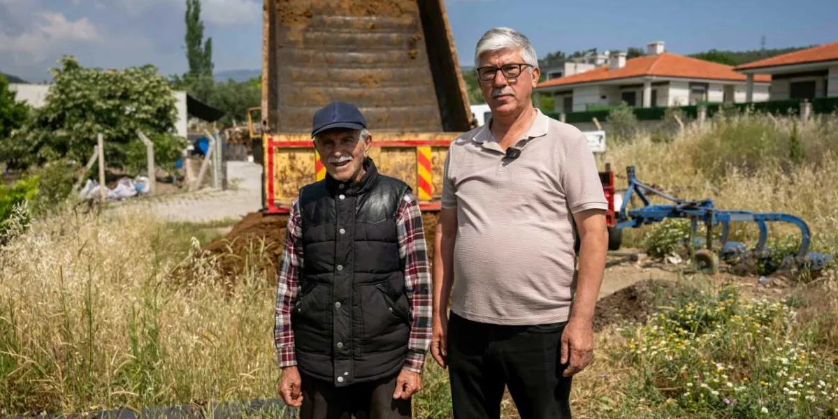 Açık havada, arkalarında kurbanlık hayvan atıklarını taşıyan sarı renkli bir damperli kamyonun bulunduğu arazide yan yana duran iki orta yaşlı erkek. Soldaki kişi lacivert şapkalı, ekose gömlekli ve siyah yelekli; sağdaki kişi ise gözlüklü ve bej renkli polo yaka tişörtlüdür. Arka planda kuru otlar, tarım aletleri ve müstakil evler görülmektedir.
