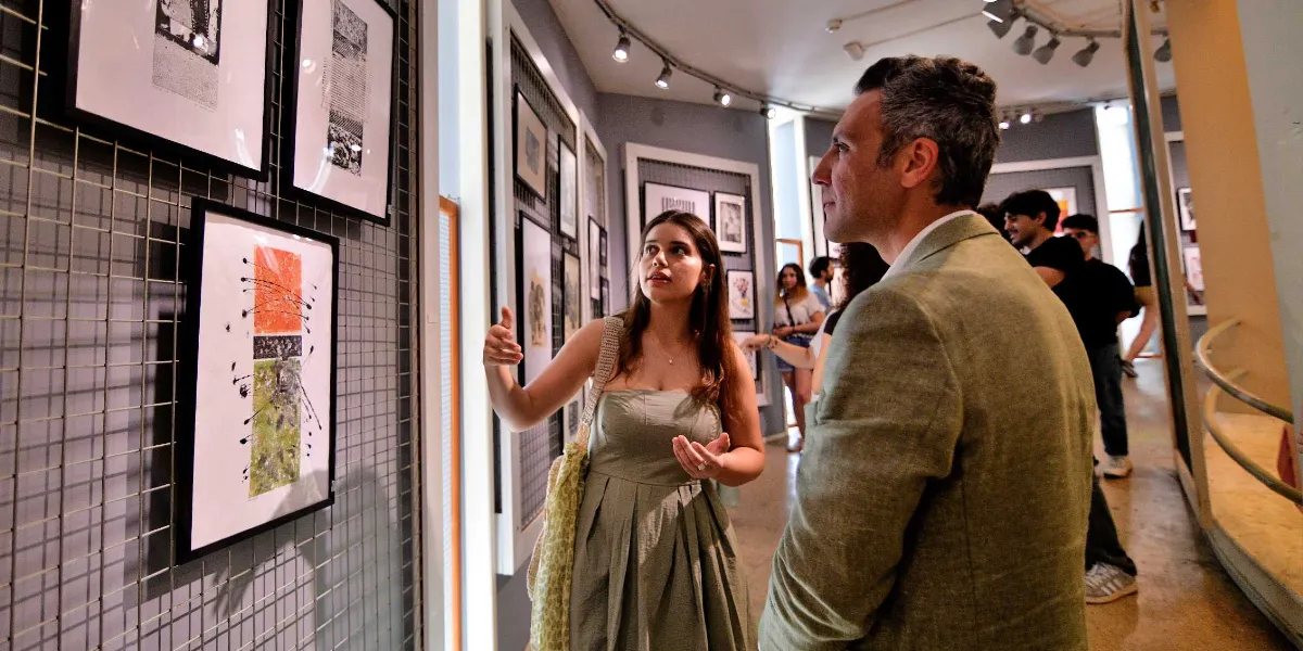 Modern bir sanat galerisinde, duvardaki metal ızgaralara asılı çerçeveli eserleri inceleyen bir kadın ve bir erkek. Uzun saçlı, askısız yeşil bir elbise giyen kadın, eliyle işaret ederek yanındaki bej ceketli erkeğe eser hakkında bilgi veriyor. Arka planda galeriyi gezen diğer ziyaretçiler ve spot ışıklarıyla aydınlatılmış sergi alanı görülüyor.