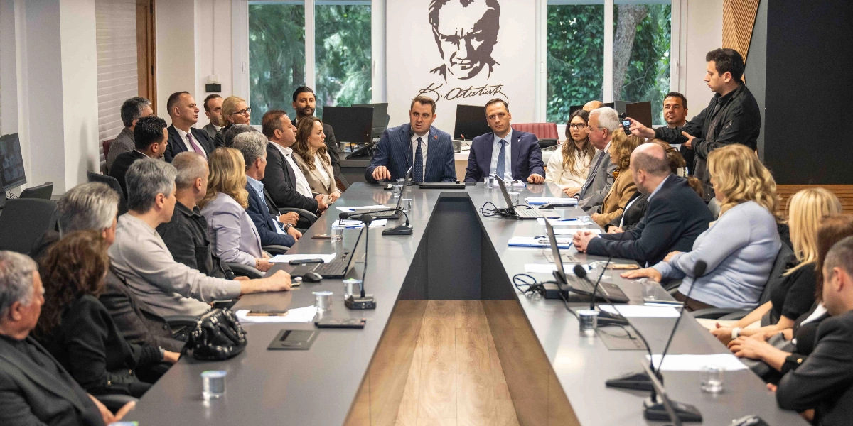 Modern bir toplantı odasında, uzun bir masa etrafında toplanmış çok sayıda kişinin katıldığı bir toplantı gerçekleşiyor. Masanın başında, arka planda duvarda Mustafa Kemal Atatürk'ün silüeti ve imzası bulunan bir tablo yer alıyor. Toplantı masasının merkezinde, takım elbiseli iki kişi konuşma yaparken, diğer katılımcılar onları dinliyor. Masanın üzerinde dizüstü bilgisayarlar, mikrofonlar ve notlar bulunuyor. Sağ tarafta elinde kayıt cihazı tutan bir kişi toplantıyı görüntülüyor. Katılımcılar resmi kıyafetler içinde, ciddi bir atmosferde bir araya gelmiş durumda.